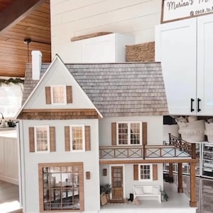 May include: A multi-story dollhouse with light gray siding, a dark shingled roof, and brown wooden trim. It features multiple windows with shutters, a front porch with a white bench, a balcony with a railing, and a chimney. The house sits on a round base.