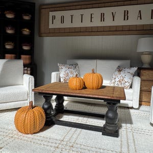 May include: Miniature interior scene with a Pottery Barn sign, a white sofa with floral pillows, and a wooden coffee table. Three orange pumpkins are on the table and floor. The room has a rug and a lamp.