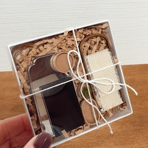 May include: A gift box containing a silver flask, a small woven rug, and other decorative items. The box is filled with brown paper and tied with white string. The flask has a black front and a silver back.