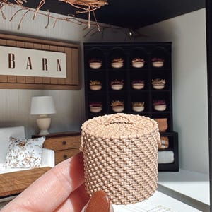 Könnte beinhalten: Ein kleiner, gewebter Korb in hellem Beige, gehalten in einer Hand. Der Korb hat ein strukturiertes, gewebtes Muster. Der Hintergrund zeigt eine Miniatur-Raumkulisse mit einem Schild mit der Aufschrift "BARN".