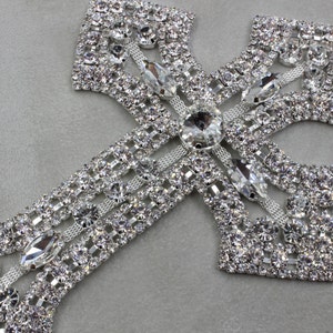 Puede incluir: Una brillante cruz plateada adornada con numerosos brillantes transparentes. La cruz presenta un diseño detallado con piedras más grandes y alargadas en el centro y en los bordes, creando un efecto deslumbrante. El fondo es gris suave.