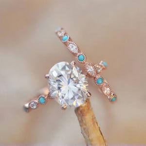 May include: Two rose gold rings. One ring features a large oval-cut diamond. The second ring has small diamonds and turquoise stones. The rings are set on a wooden stick against a blurred background.