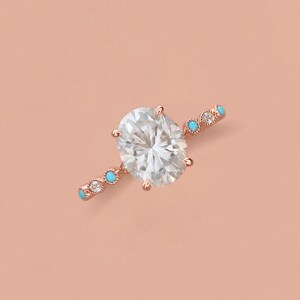 May include: A rose gold ring featuring a large oval-cut clear center stone. The band is accented with alternating small round clear stones and small round turquoise-colored stones set in a delicate metal frame with a beaded texture.