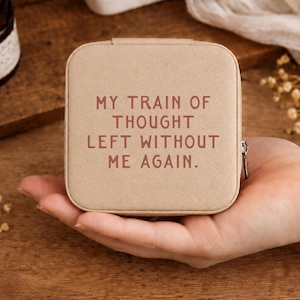 May include: A square, beige jewelry travel case with a textured finish. The top of the case features the text in dark brown letters: MY TRAIN OF THOUGHT LEFT WITHOUT ME AGAIN. The case has a silver-toned metal zipper pull on the right side.