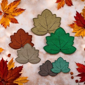 May include: Five felt leaf-shaped coasters in various colours, including brown, green, and beige. The coasters are arranged on a light brown background.