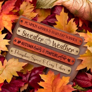 May include: Four fabric wristbands with autumn-themed text. The top band is red and says "Pumpkin Kisses & Harvest Wishes". The second band is gold and says "Sweater Weather" with a pumpkin icon. The third band is red and says "Autumn Leaves & Pumpkins Please". The bottom band is gold and says "Pumpkin Spice & Cozy Vibes".