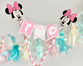 Op de afbeelding: Een taarttopper met een roze en witte banner met de tekst "Two", geflankeerd door Minnie Mouse-hoofden met roze strikken. De topper wordt ondersteund door roze en wit gestreepte rietjes. Pastelkleurige decoraties op de achtergrond.