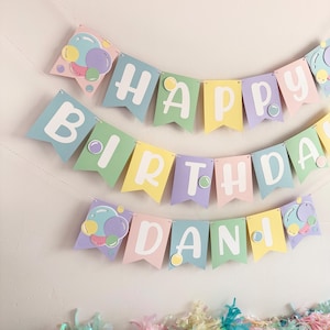May include: A pastel-colored birthday banner with the words "HAPPY BIRTHDAY DANI" in white letters. The banner is decorated with bubble-shaped accents and a colorful tassel garland.