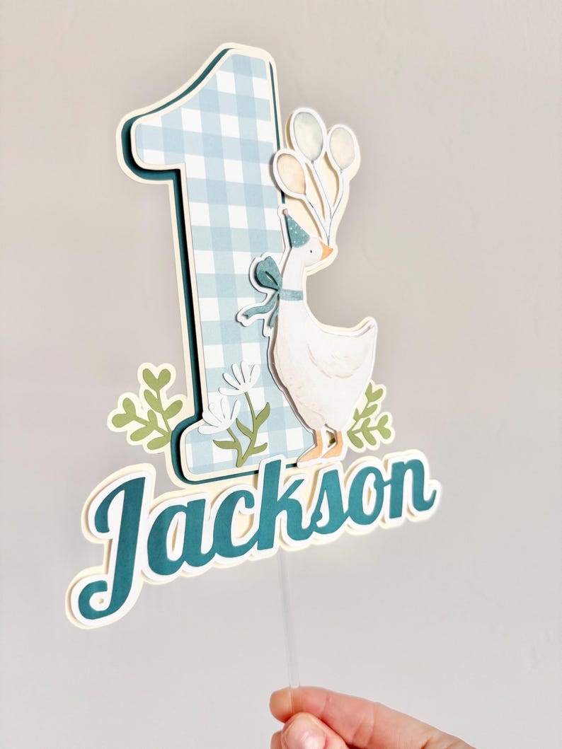 Pu&ograve; includere: Decorazione per torta con il numero uno a quadretti blu e bianchi, con un'oca dei cartoni animati che tiene in mano dei palloncini. Il nome "Jackson" &egrave; scritto in corsivo verde acqua. Il design include foglie verdi e fiori bianchi.