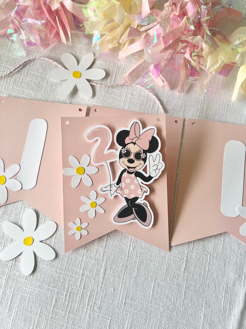 Puede incluir: Una pancarta rosa y blanca con una imagen de dibujos animados de un rat&oacute;n con gafas de sol y sosteniendo un globo. La pancarta tiene el n&uacute;mero "2" y est&aacute; decorada con margaritas blancas.