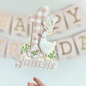 Pu&ograve; includere: Un cake topper personalizzato per un primo compleanno. Presenta il numero uno con un motivo a quadretti rosa, un'oca bianca con un cappello da festa e il nome "Juliette". Decorato con verde e palloncini.