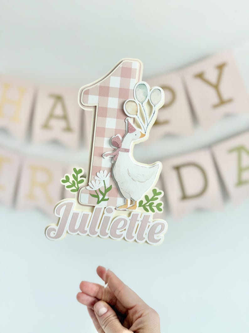 Pu&ograve; includere: Un cake topper personalizzato per un primo compleanno. Presenta il numero uno con un motivo a quadretti rosa, un'oca bianca con un cappello da festa e il nome "Juliette". Decorato con verde e palloncini.