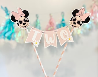 May include: A pink and white cake topper featuring two Minnie Mouse heads, each with sunglasses and a pink bow. The banner spells "TWO" and is decorated with daisies. The topper is supported by two pink and white striped straws.