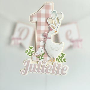 Pu&ograve; includere: Un cake topper con un numero uno rosa a quadretti e un'oca bianca con un cappello da festa e palloncini. Il nome "Juliette" &egrave; scritto in corsivo sotto.