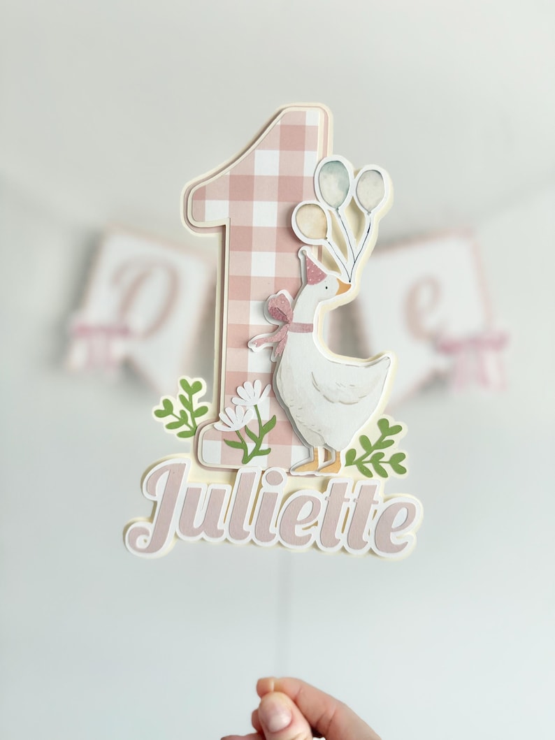 Pu&ograve; includere: Un cake topper con un numero uno rosa a quadretti e un'oca bianca con un cappello da festa e palloncini. Il nome "Juliette" &egrave; scritto in corsivo sotto.
