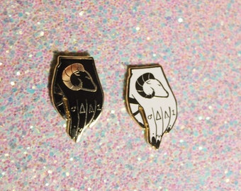 May include: Two enamel pins, one black and one white, each depicting a hand holding a ram's head. The pins have gold accents and the text "ΔΔN1" on the fingers.