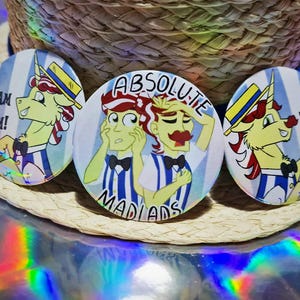 May include: Three circular badges featuring cartoon characters. Two badges display a unicorn in a hat with the text "TEAM FLIM!" and "TEAM FLAM!". The central badge shows two men with the text "ABSOLUTE MADIADS!".