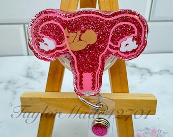 May include: Pink glitter badge reel with a pink and white outline of a uterus with a baby inside. The reel has a silver swivel clip and a pink glitter charm.