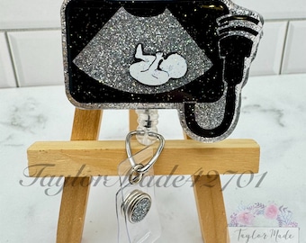 May include: A black and silver glitter badge reel with a white silhouette of a baby in an ultrasound image. The reel has a silver glitter retractable cord and a silver glitter badge clip.