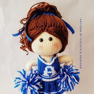 Pode incluir: Boneca de malha vestindo um traje de líder de torcida azul e branco com pompons. A boneca tem cabelo castanho em um rabo de cavalo e olhos azuis. A boneca está em pé em um fundo branco. PATTERN BY VIOLETS & HEATHER