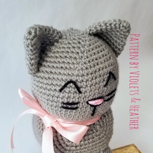 May include: A gray crocheted cat with a pink bow, sitting on a vintage book with a bird illustration. The cat has black embroidered eyes and a pink nose.