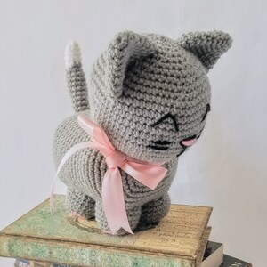 May include: A gray crocheted cat toy with a pink bow. The cat is sitting on a stack of books.