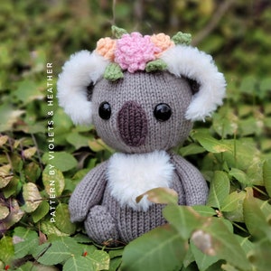 May include: A gray knitted koala with white fur on its ears and chest, wearing a flower crown with pink, orange, and green flowers. The koala is sitting in a bed of green leaves.