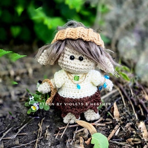 Peut inclure: Une poupée gnome en crochet avec des cheveux bruns, portant une chemise blanche, une jupe brune et un chapeau en crochet beige. Le gnome tient un petit panier et se tient sur un lit de feuilles et de brindilles brunes. Le texte "PATTERN BY VIOLETS & HEATHER" est visible sur l'image.