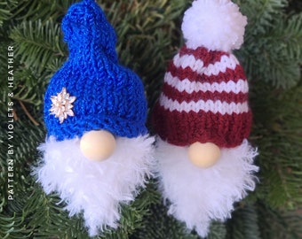 May include: Two knitted gnome ornaments hanging on a green Christmas tree. One gnome is wearing a blue hat with a white pom-pom and a white beard. The other gnome is wearing a red and white striped hat with a white pom-pom and a white beard.