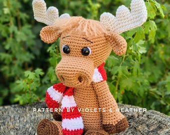 Puede incluir: Un alce de crochet marrón con cuernos blancos y una bufanda a rayas rojas y blancas. El alce está sentado en un tocón de árbol. El texto "PATTERN BY VIOLETS & HEATHER" es visible en el tocón.