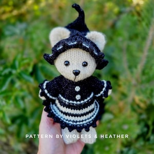 Op de afbeelding: Een handgemaakte gebreide beer met een zwart-witte outfit, inclusief een hoed en schort. De beer heeft een beige gezicht met zwarte ogen en een zwarte neus. De tekst "PATTERN BY VIOLETS & HEATHER" staat onderaan.
