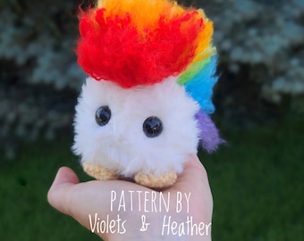May include: A white crocheted plushie with a rainbow mane and a red mohawk. The plushie has large black eyes and is being held in a hand. The text "PATTERN BY Violets & Heather" is visible below the plushie.
