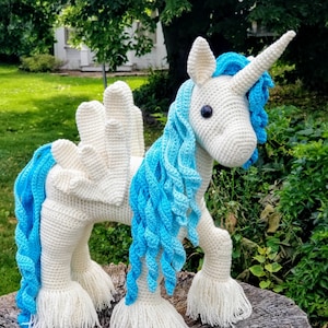 May include: A white crocheted unicorn with blue yarn mane and tail. The unicorn has a white crocheted horn and black eyes. The unicorn is standing on a tree stump.