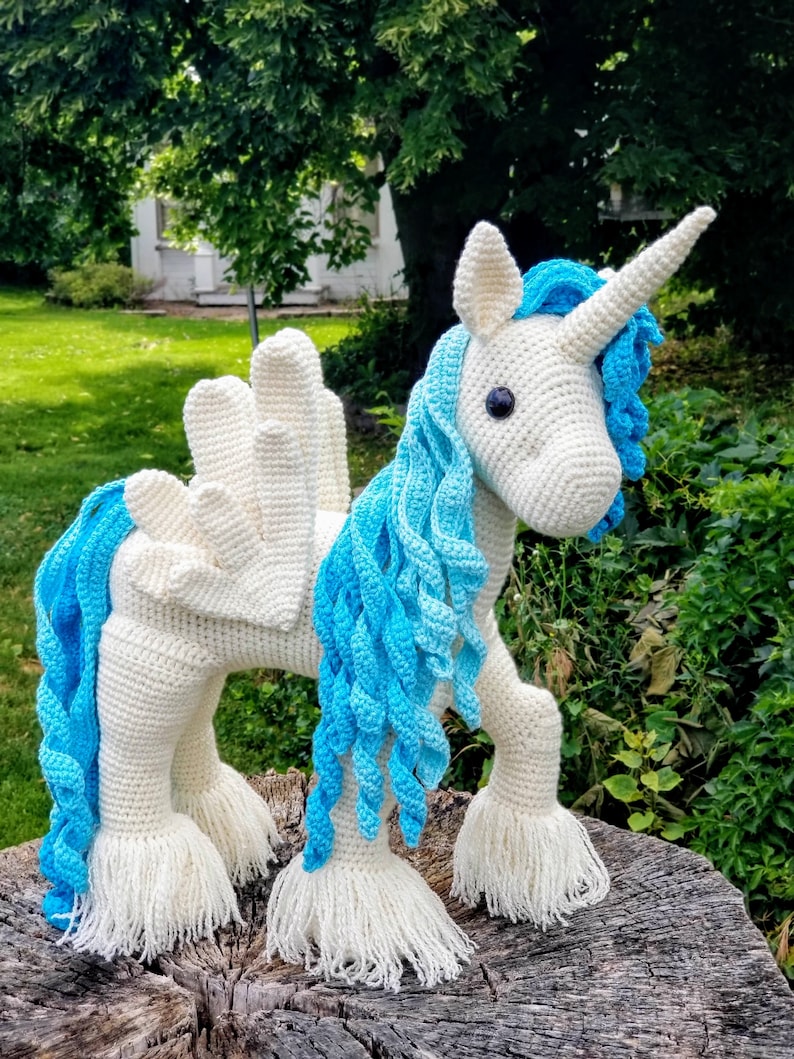 May include: A white crocheted unicorn with blue yarn mane and tail. The unicorn has a white crocheted horn and black eyes. The unicorn is standing on a tree stump.
