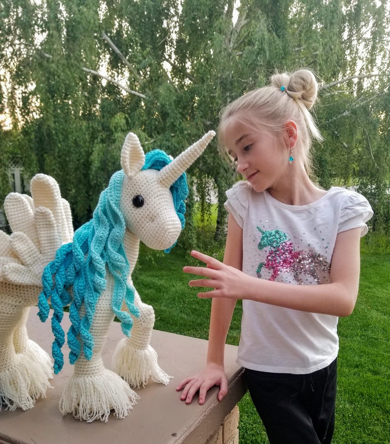 May include: A white crocheted unicorn with blue yarn mane and tail. A young girl is looking at the unicorn with her hand outstretched.
