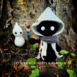 May include: Two crocheted dolls, one white and one black, stand in front of a tree. The black doll wears a gray scarf and a light gray hooded cloak. The white doll is smaller and has a simple design. A wooden staff with beads is between them. The text "PATTERN BY VIOLETS & HEATHER" is at the bottom.