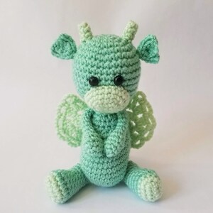May include: A crocheted green dragon with wings, sitting with its arms crossed. The dragon has black eyes and a light green face.