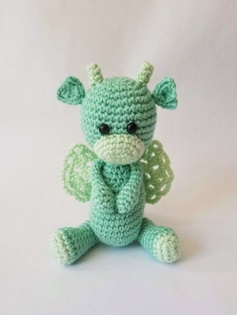 May include: A crocheted green dragon with wings, sitting with its arms crossed. The dragon has black eyes and a light green face.