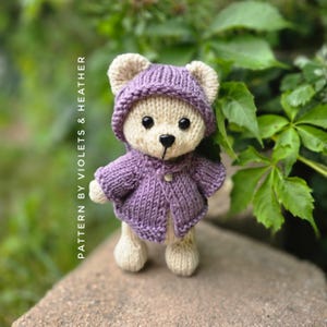 May include: A handmade knitted teddy bear wearing a purple hooded jacket. The bear has a cream-colored body, beige ears, and a black nose. The text "PATTERN BY VIOLETS & HEATHER" is printed vertically on the left side of the image.