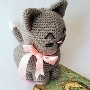 May include: A gray crocheted cat toy with a pink ribbon around its neck. The cat has black embroidered eyes and a pink nose. The cat is sitting on a vintage book with a bird illustration.