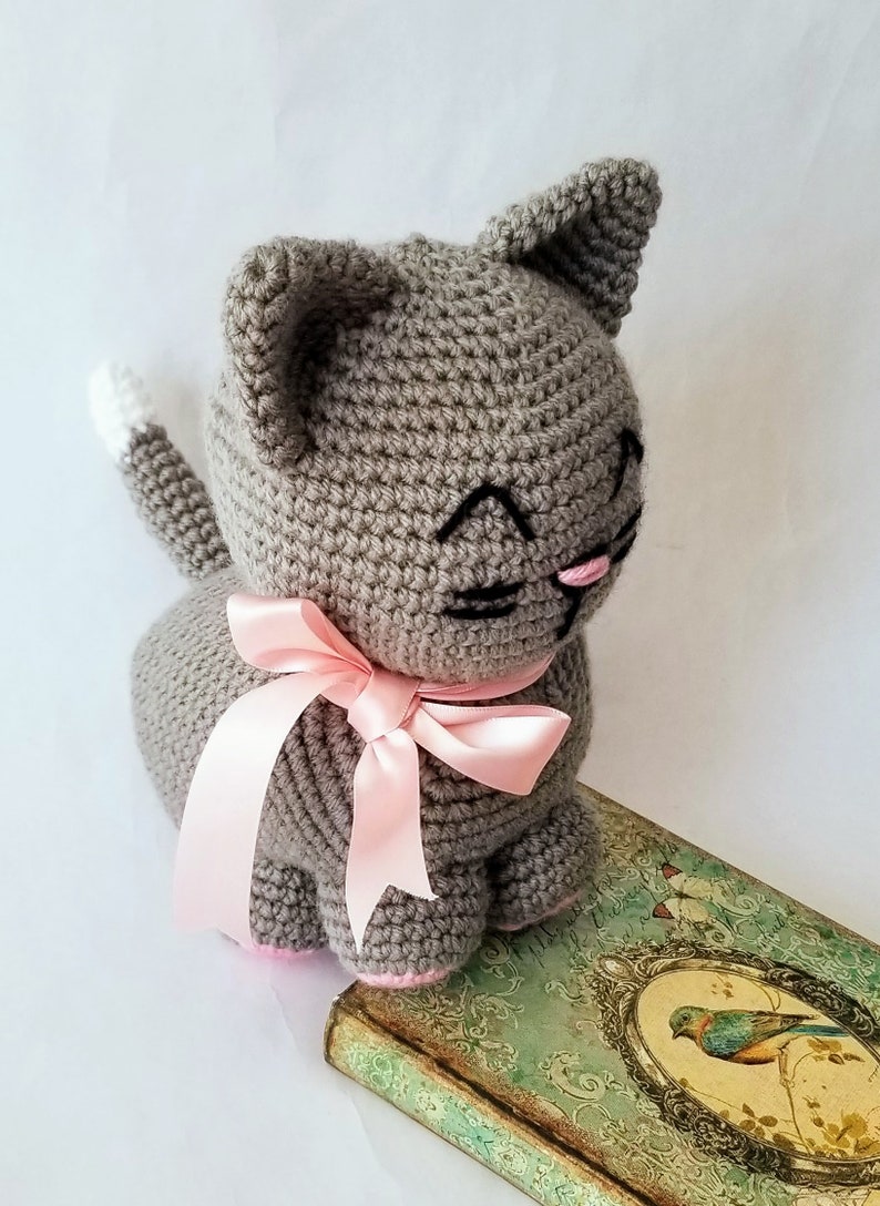 May include: A gray crocheted cat toy with a pink ribbon around its neck. The cat has black embroidered eyes and a pink nose. The cat is sitting on a vintage book with a bird illustration.