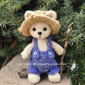 May include: A handmade knitted teddy bear wearing a tan hat and blue overalls with button details. The bear is holding a small stick and is posed in front of green foliage. The text "PATTERN BY VIOLETS & HEATHER" is visible.