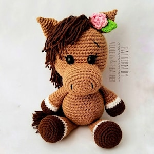 May include: A brown crocheted horse toy with a pink flower and green leaf on its head. The horse has brown eyes, a brown tail, and white and brown hooves. The toy is sitting on a white background. "PATTERN BY VIOLETS & HEATHER" is written on the background.