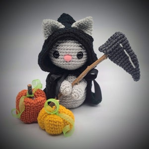 May include: A gray crocheted cat dressed as the Grim Reaper holding a crocheted scythe and standing next to two crocheted pumpkins, one orange and one yellow.