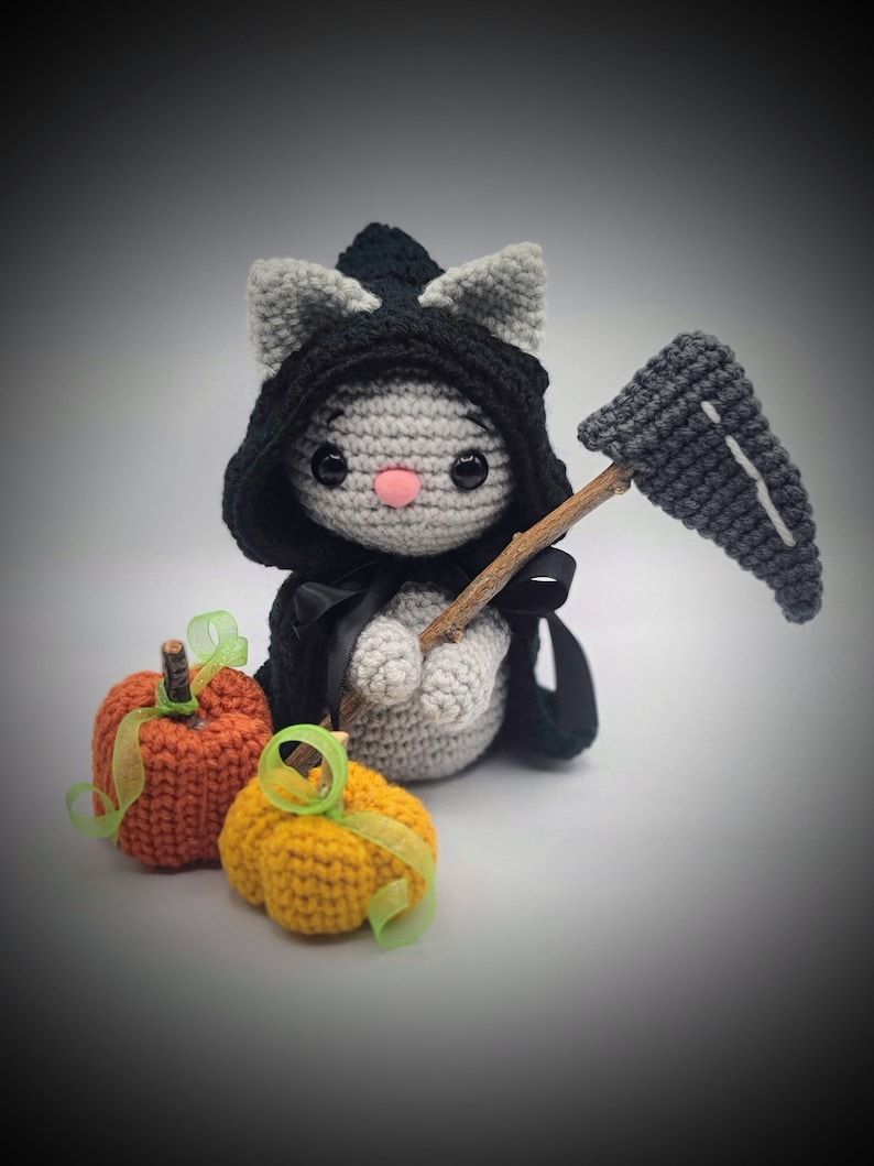May include: A gray crocheted cat dressed as the Grim Reaper holding a crocheted scythe and standing next to two crocheted pumpkins, one orange and one yellow.
