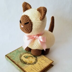 May include: A crocheted cat toy with brown ears, brown eyes, and a pink bow. The cat is sitting on a vintage book with a bird illustration.
