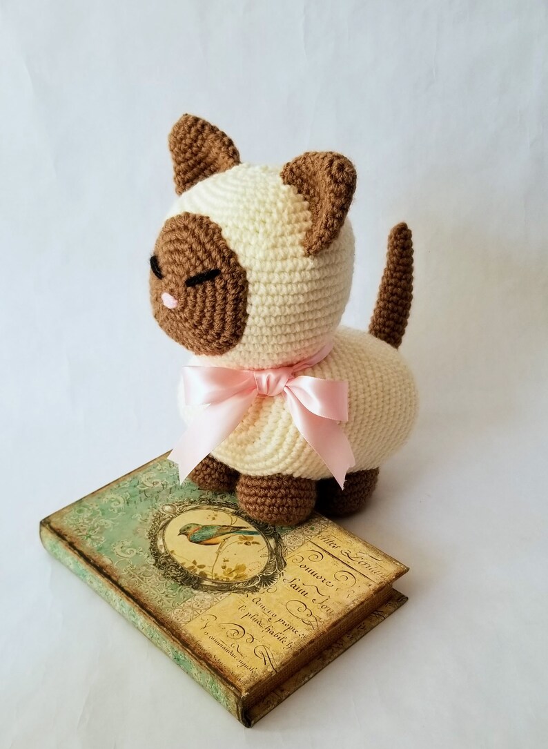 May include: A crocheted cat toy with brown ears, brown eyes, and a pink bow. The cat is sitting on a vintage book with a bird illustration.