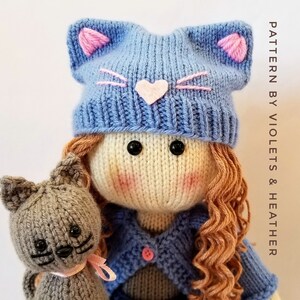 May include: A knitted doll with long brown hair wearing a blue knitted hat with pink cat ears and a blue knitted cardigan. The doll is holding a grey knitted cat with black eyes.