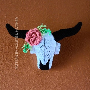 May include: White crocheted cow skull with black horns and a pink flower with green leaves.