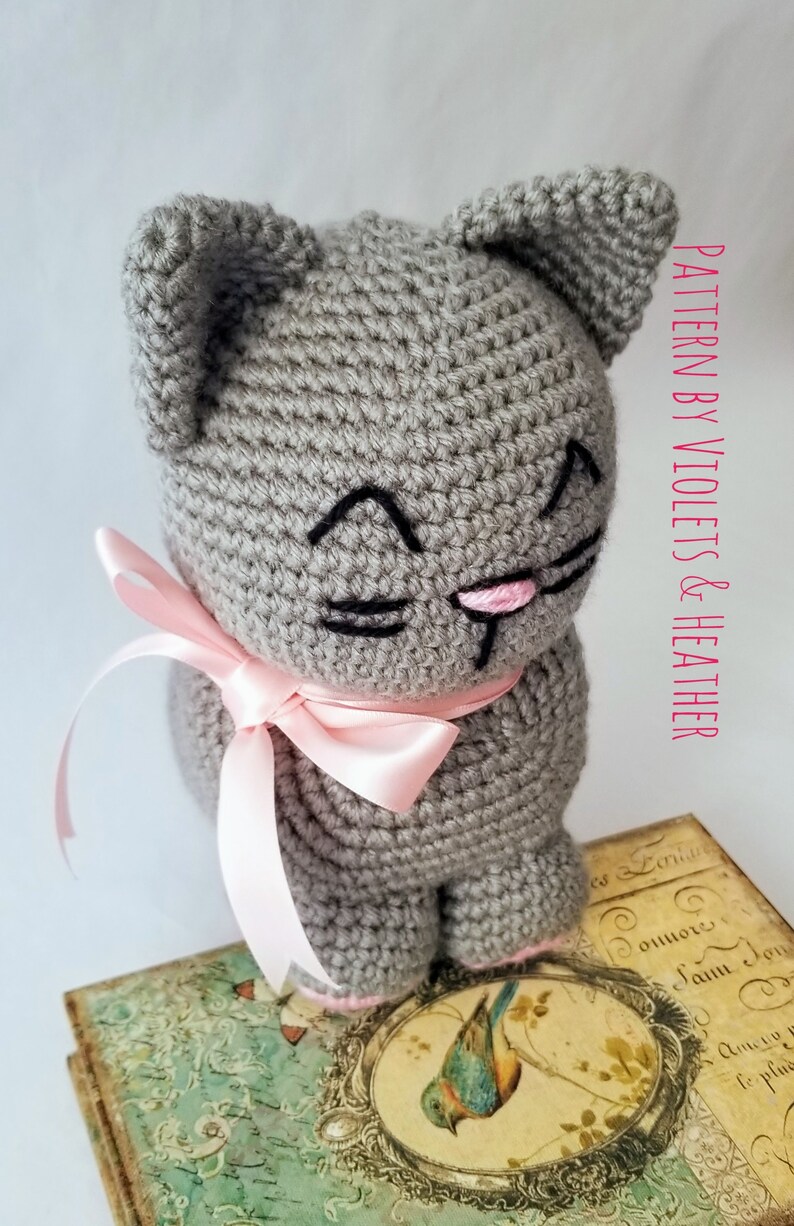 May include: A gray crocheted cat with a pink bow, sitting on a vintage book with a bird illustration. The cat has black embroidered eyes and a pink nose.